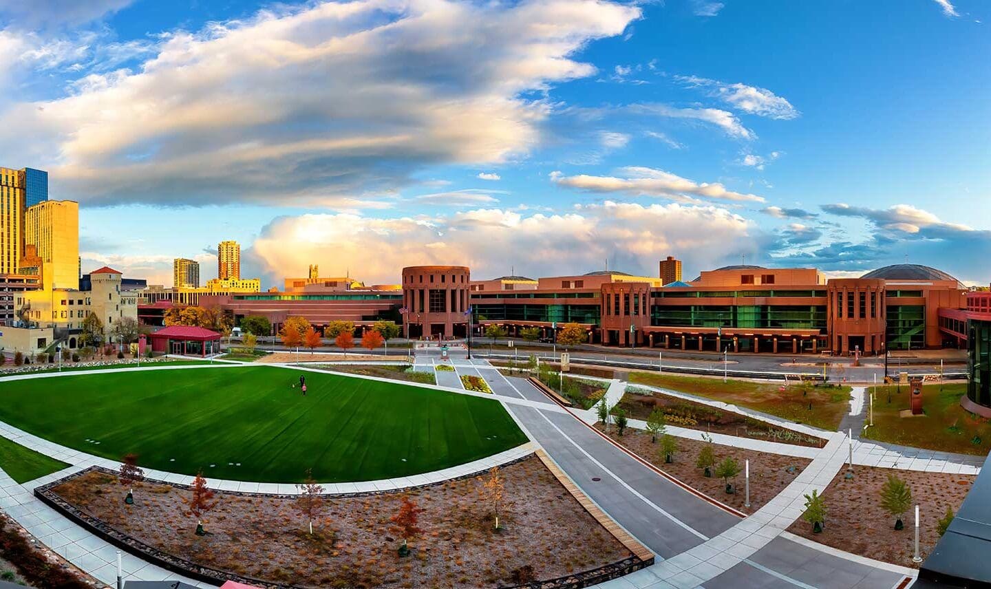 A panoramic view of a modern urban campus near the Minneapolis Convention Center, with red brick buildings, green lawn, walkways, and a partly cloudy sky at sunset.