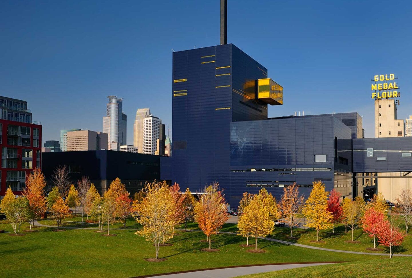 A modern dark blue building, the Guthrie Theater, stands behind a row of colorful autumn trees, with city skyscrapers and a Gold Medal Flour sign visible in the background.