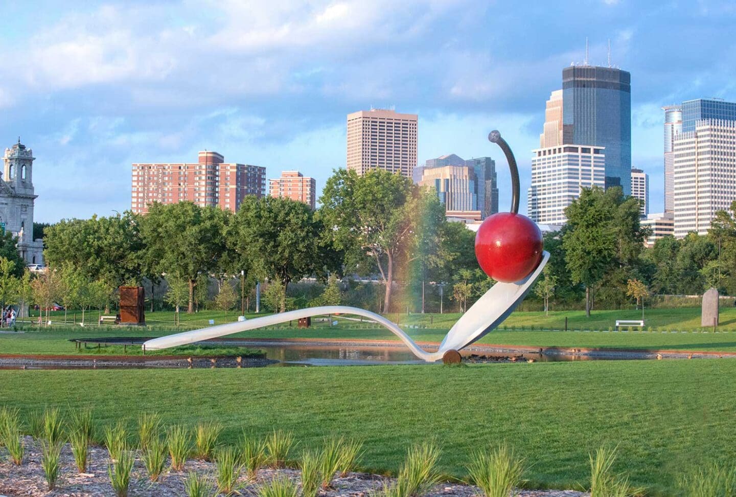 Large outdoor sculpture of a spoon with a cherry on it, set in the Sculpture Garden at Walker Art Center, surrounded by grassy parkland with city buildings in the background.