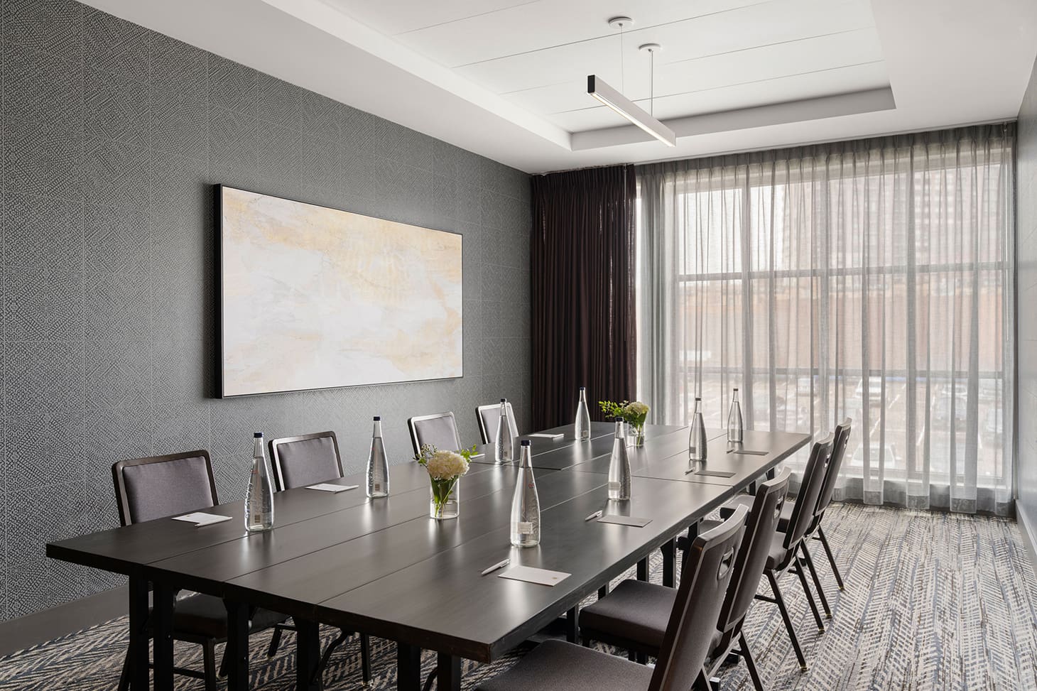 A modern conference room with a long table, eight chairs, bottled water, notepads, small flower vases, large window with sheer curtains, and abstract art on the wall.