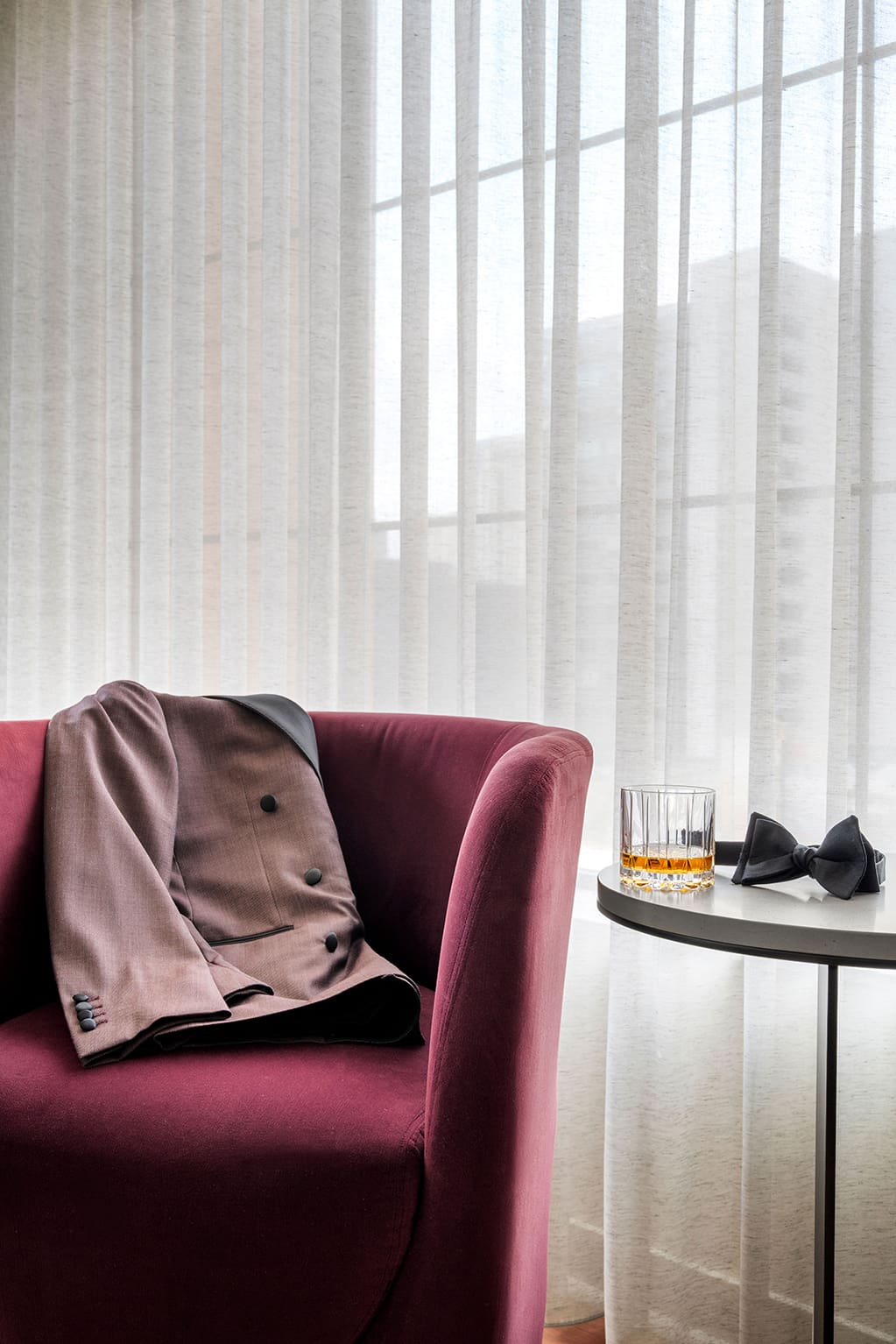 A mauve suit jacket draped over a burgundy chair next to a round table holding a glass of whiskey and a black bow tie, set against sheer curtains with a sweeping city view in the background—evoking the refined atmosphere of a penthouse hotel room in Minneapolis.