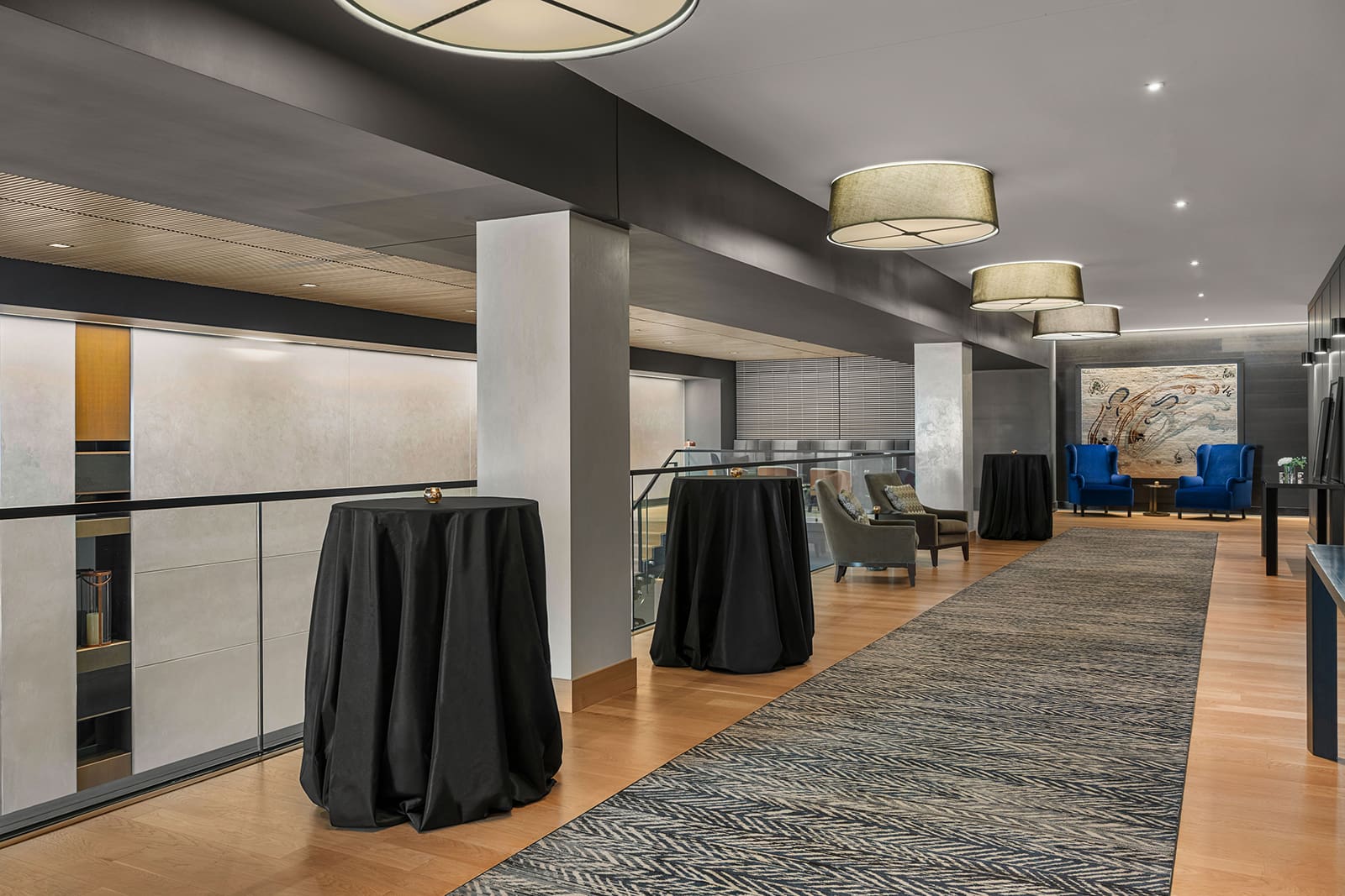 Modern pre-function space featuring a hotel hallway with wooden floors, round ceiling lights, cocktail tables covered in black cloths, lounge chairs, and a large artwork in the background.
