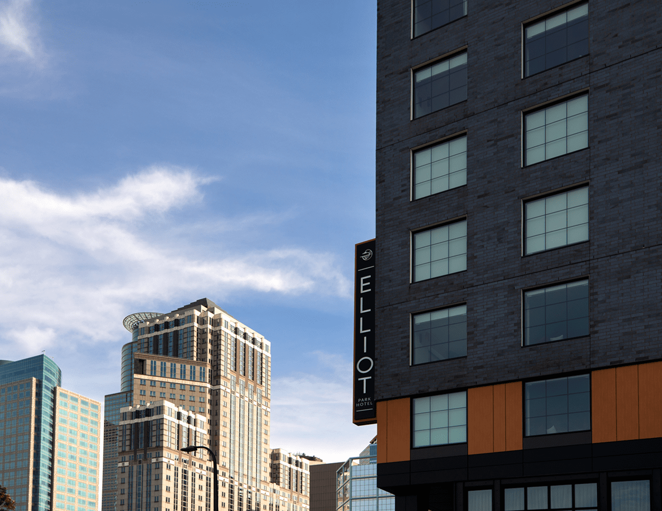 Modern high-rise buildings against a blue sky with scattered clouds; a sign on the foreground building reads Elliot.