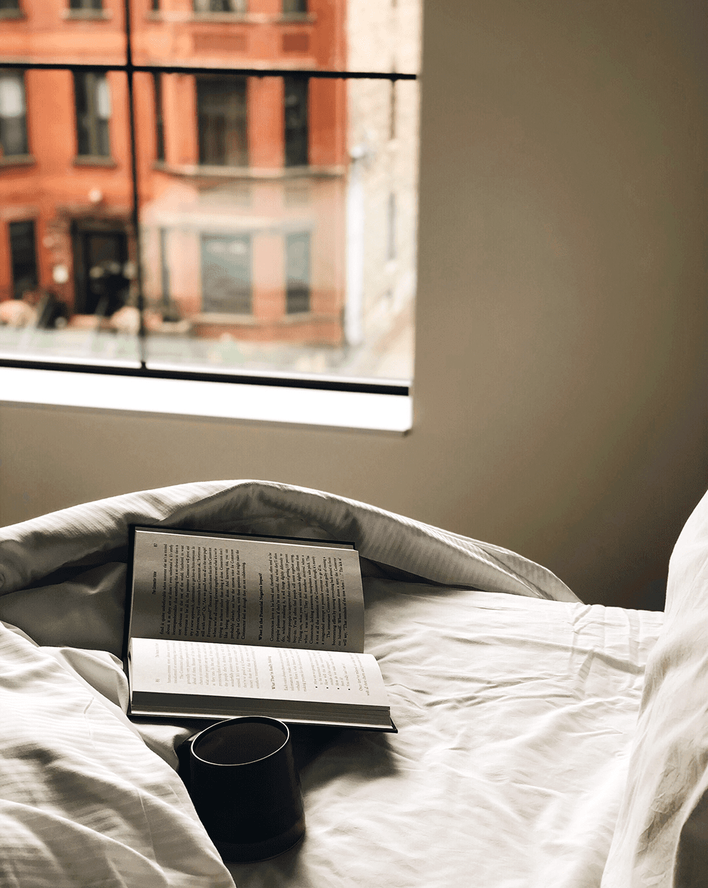 An open book and a mug rest on a bed near a window, with a view of red brick buildings outside.