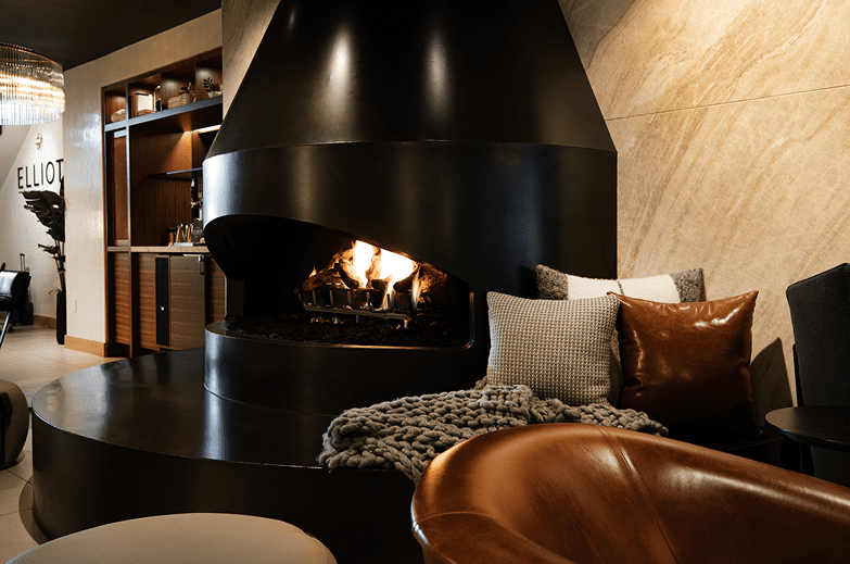 A modern lounge area with a large black fireplace, a burning fire, cushioned seating, and a blanket draped over a sofa.