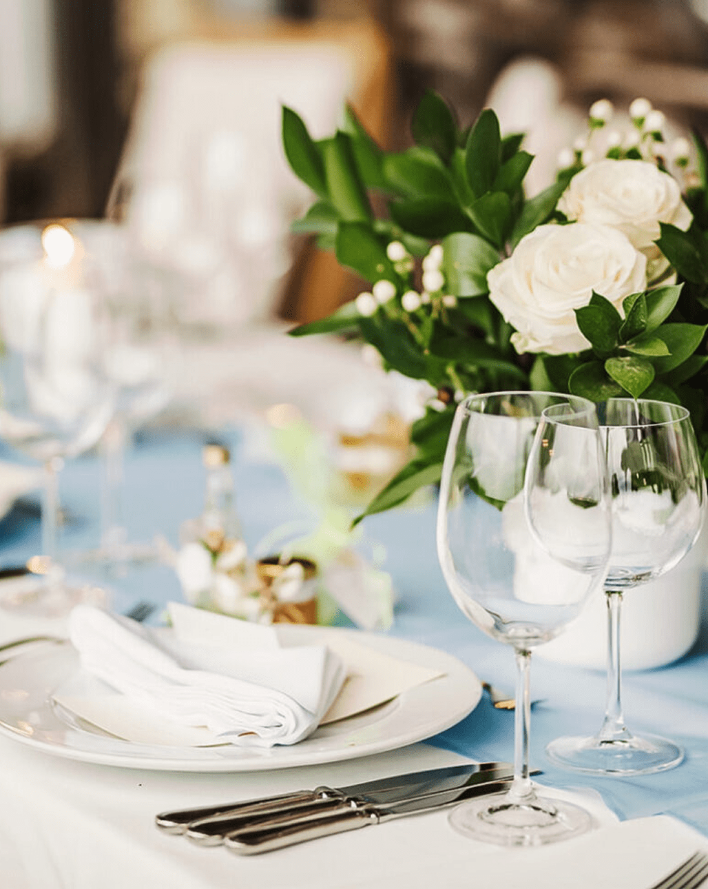 A formal dining table set with plates, cutlery, wine glasses, a white napkin, and a floral centerpiece with white roses and green leaves on a blue tablecloth.