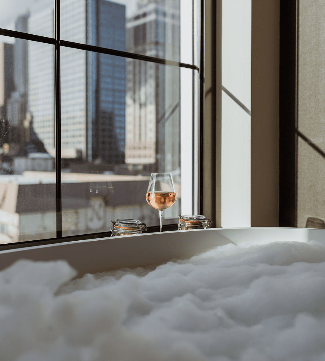 A glass of rosé sits on a windowsill next to a bubble-filled bathtub, with tall city buildings visible through the window in daylight.
