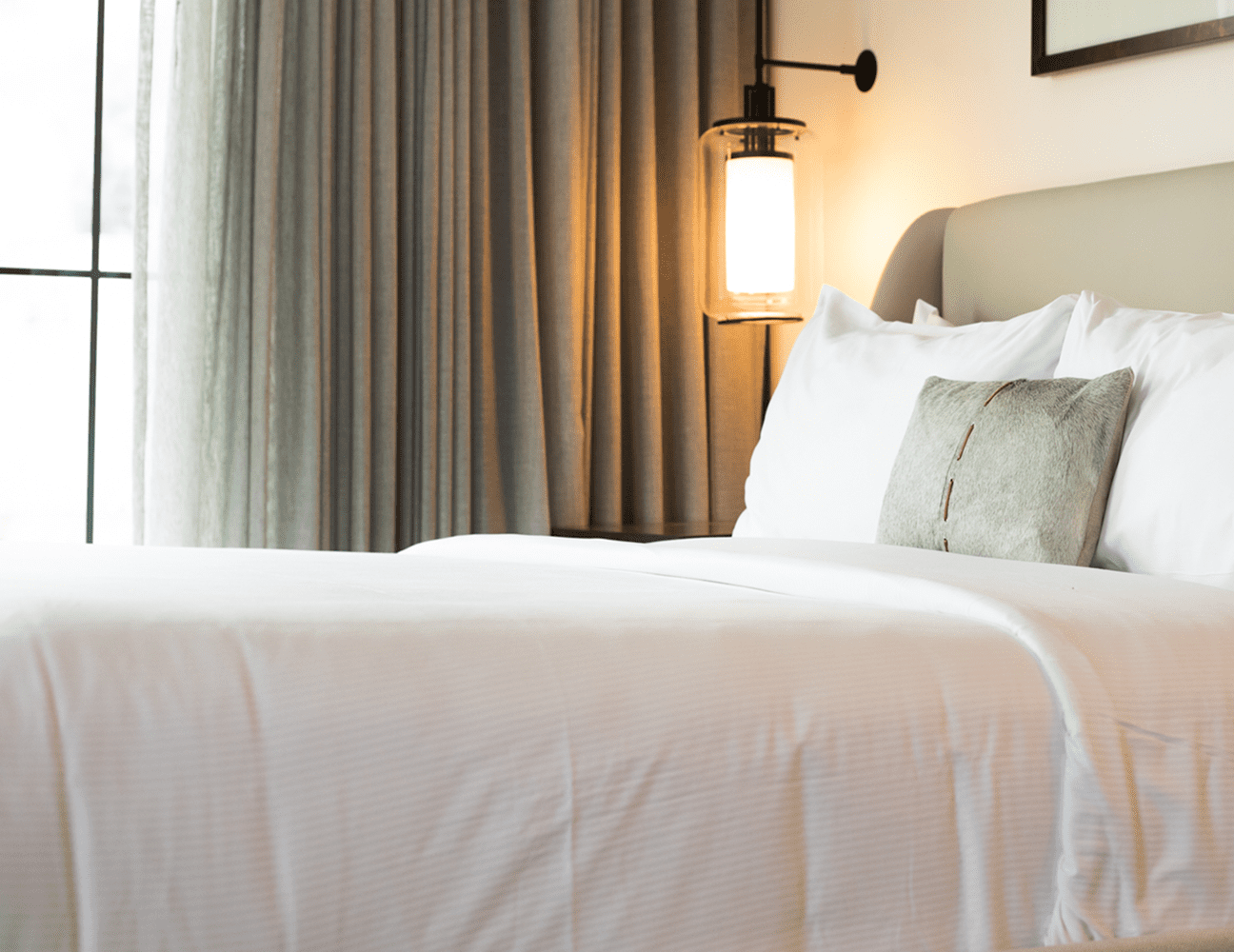 A neatly made bed with white linens and a decorative pillow, beside a lit wall lamp and floor-to-ceiling curtains.