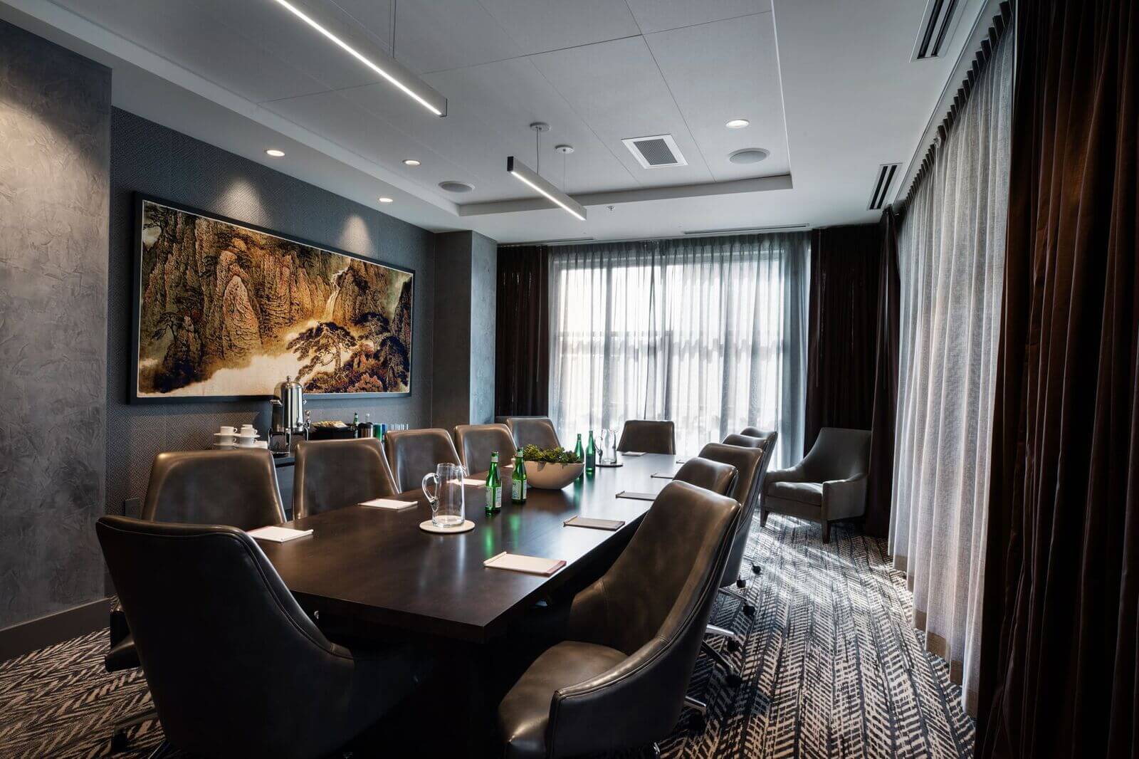 The Blaze Boardroom is a modern conference room featuring a long table, leather chairs, notepads, water bottles, glassware, and a large window with sheer curtains. A landscape painting hangs on the wall.