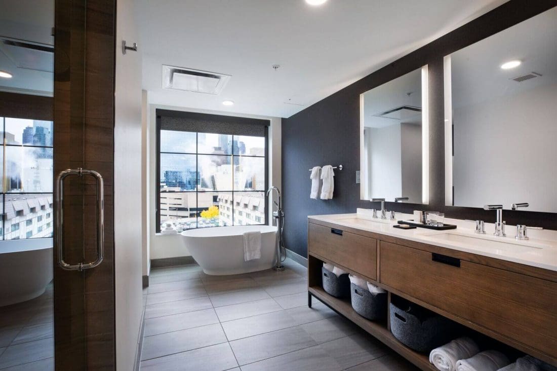 Modern bathroom with a large mirror, double sink vanity, freestanding bathtub by the window, and city buildings visible outside.