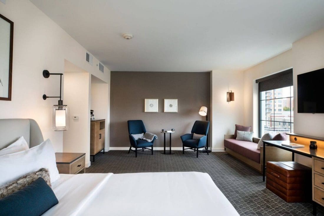 Modern hotel room with a neatly made bed, two chairs and a table, a window seat, a desk with a TV, and neutral-colored decor.