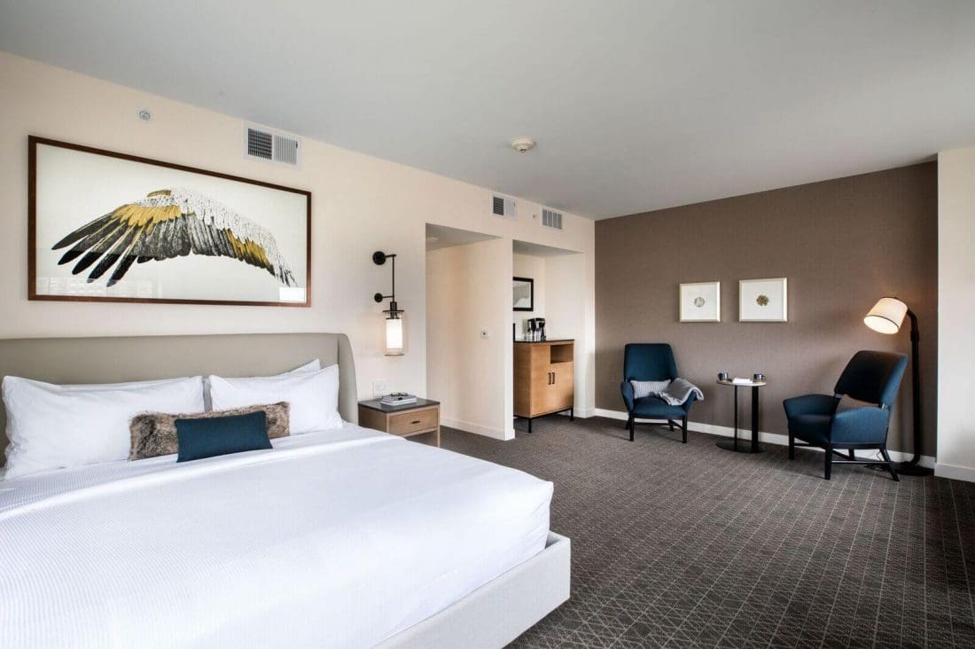 Modern hotel room with a large bed, framed bird artwork above, sitting area with two blue chairs and a table, neutral colors, and minimal decor.