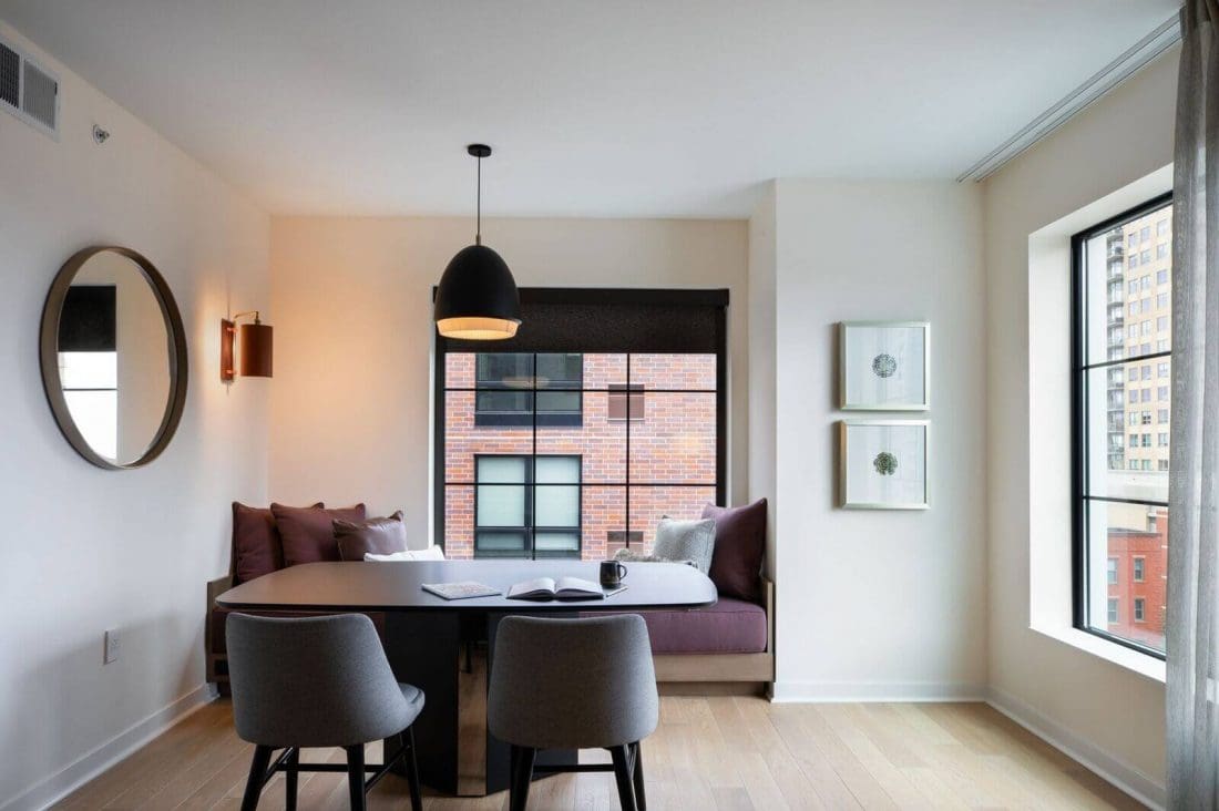 A modern dining area with a black table, two gray chairs, a built-in bench with cushions, large windows, and minimal wall decor in a bright apartment.