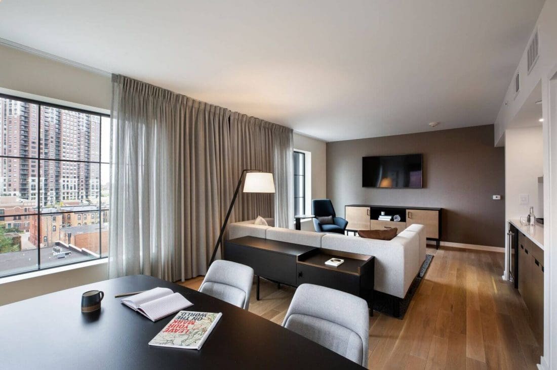 Modern living room with wood flooring, a sectional sofa, TV on the wall, large windows with city view, and a table with notebooks, a magazine, and a cup.