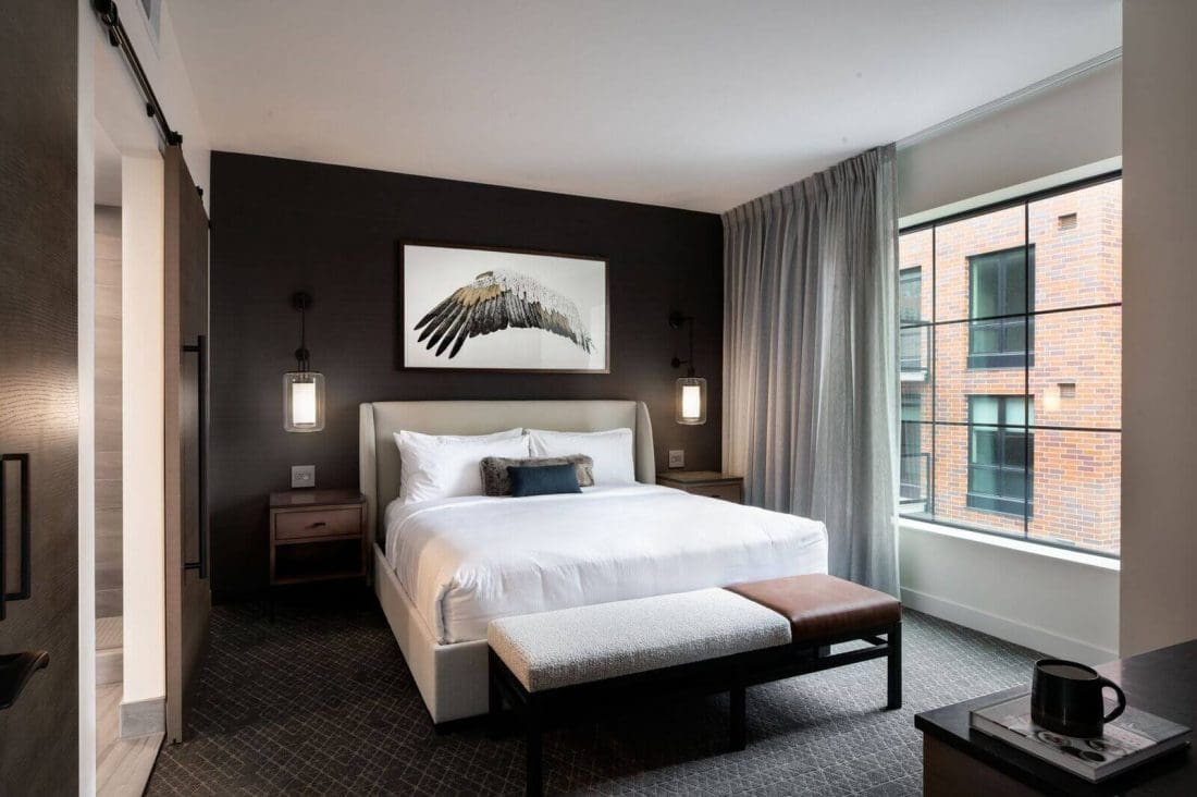 Modern hotel room with a large bed, two nightstands, a bench, wall art of a bird wing, floor-to-ceiling window with curtains, and a view of a brick building outside.