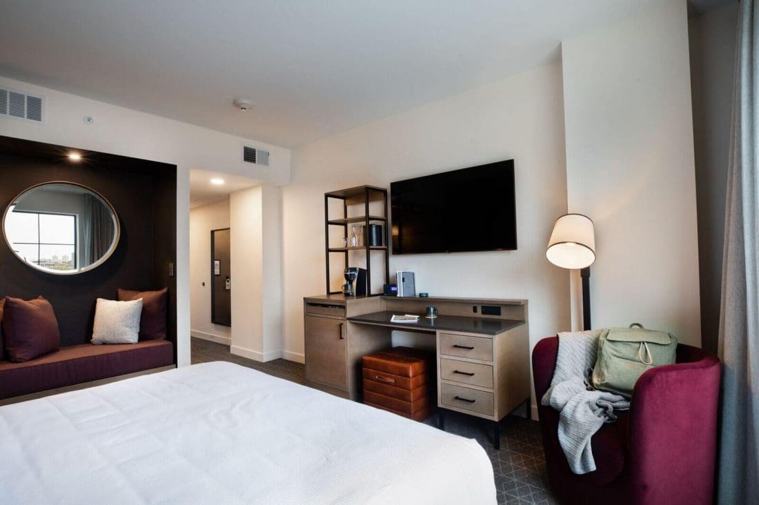 Modern hotel room with a neatly made bed, wall-mounted TV, desk with chair, round mirror, cushioned seating area, and a window with natural light.