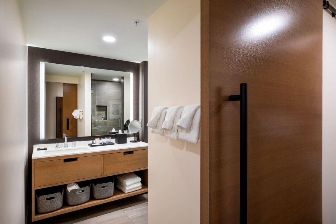 Modern bathroom with a large mirror, double sink vanity, rolled towels on the wall, and open shelving with baskets and folded towels; glass shower visible in the background.