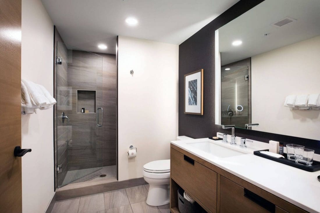 Modern bathroom in the Deluxe Queen features a glass-enclosed shower, toilet, large mirror, wooden vanity with double sinks, and towels neatly arranged on racks.