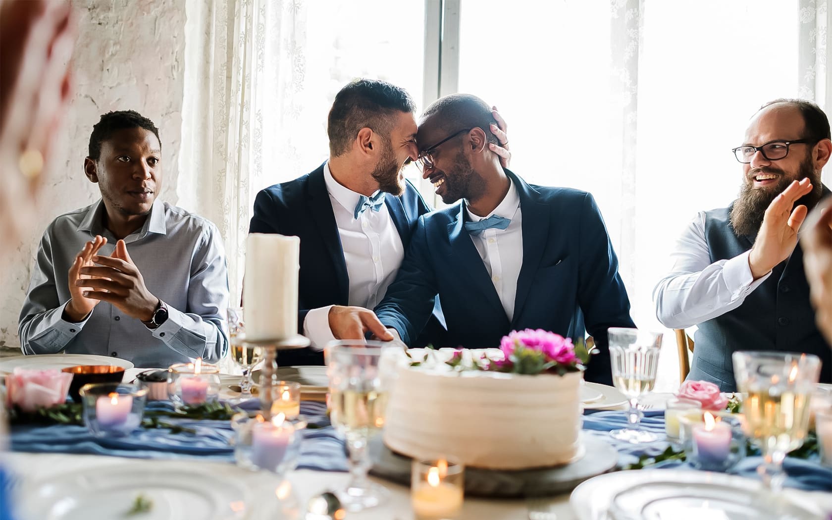 Two men in suits sit closely, smiling and touching foreheads at a decorated table with cake, candles, and friends applauding or smiling around them.