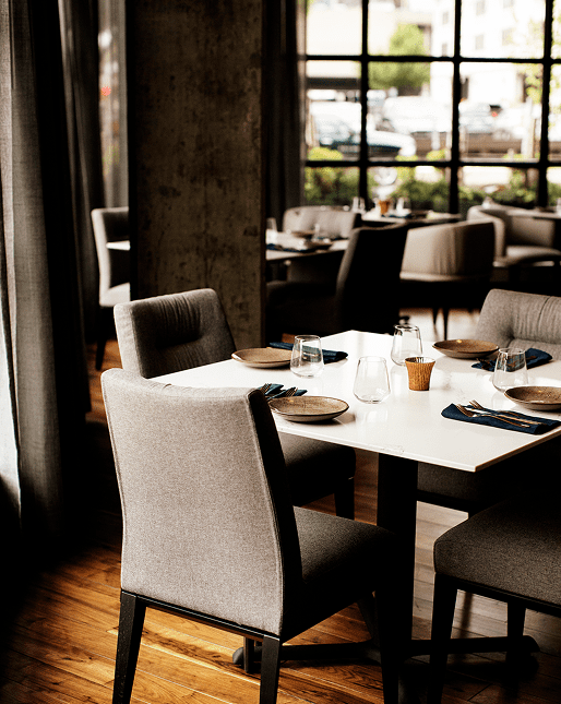 A modern restaurant dining area with neatly set tables, gray upholstered chairs, and large windows letting in natural light.