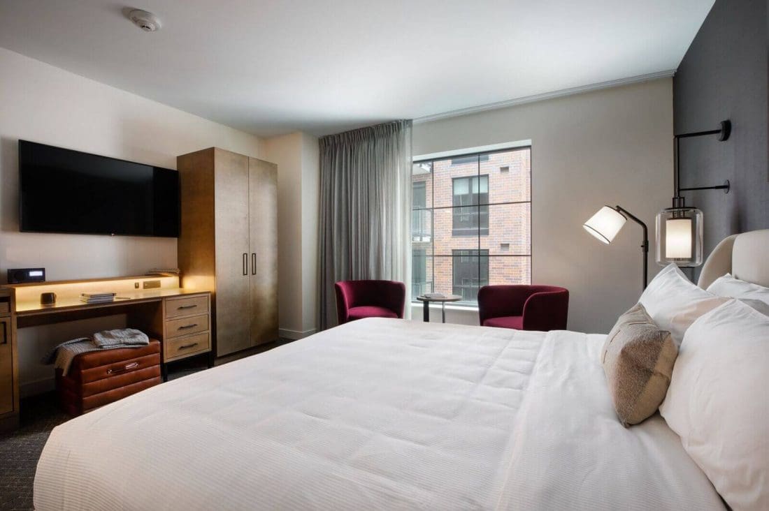 Modern hotel room with a large bed, two red chairs by a window, a desk with a TV above it, and a wardrobe against the wall.