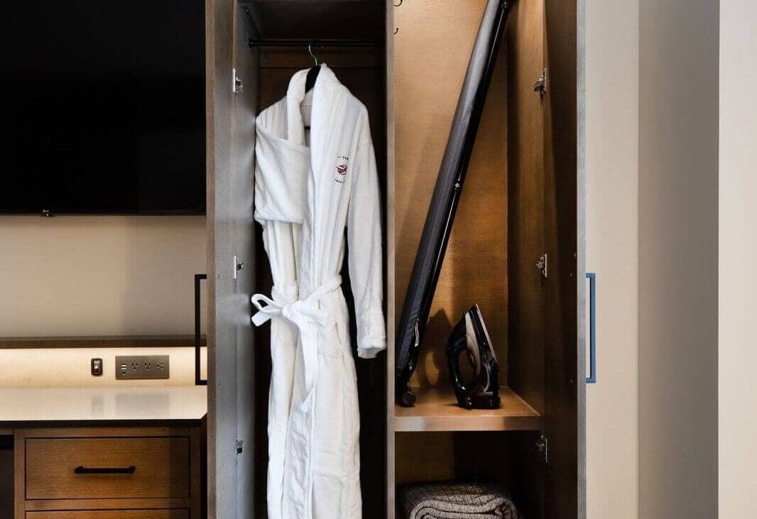 Open wardrobe with a white bathrobe on a hanger, an ironing board, an iron, and a folded gray blanket on the shelf.