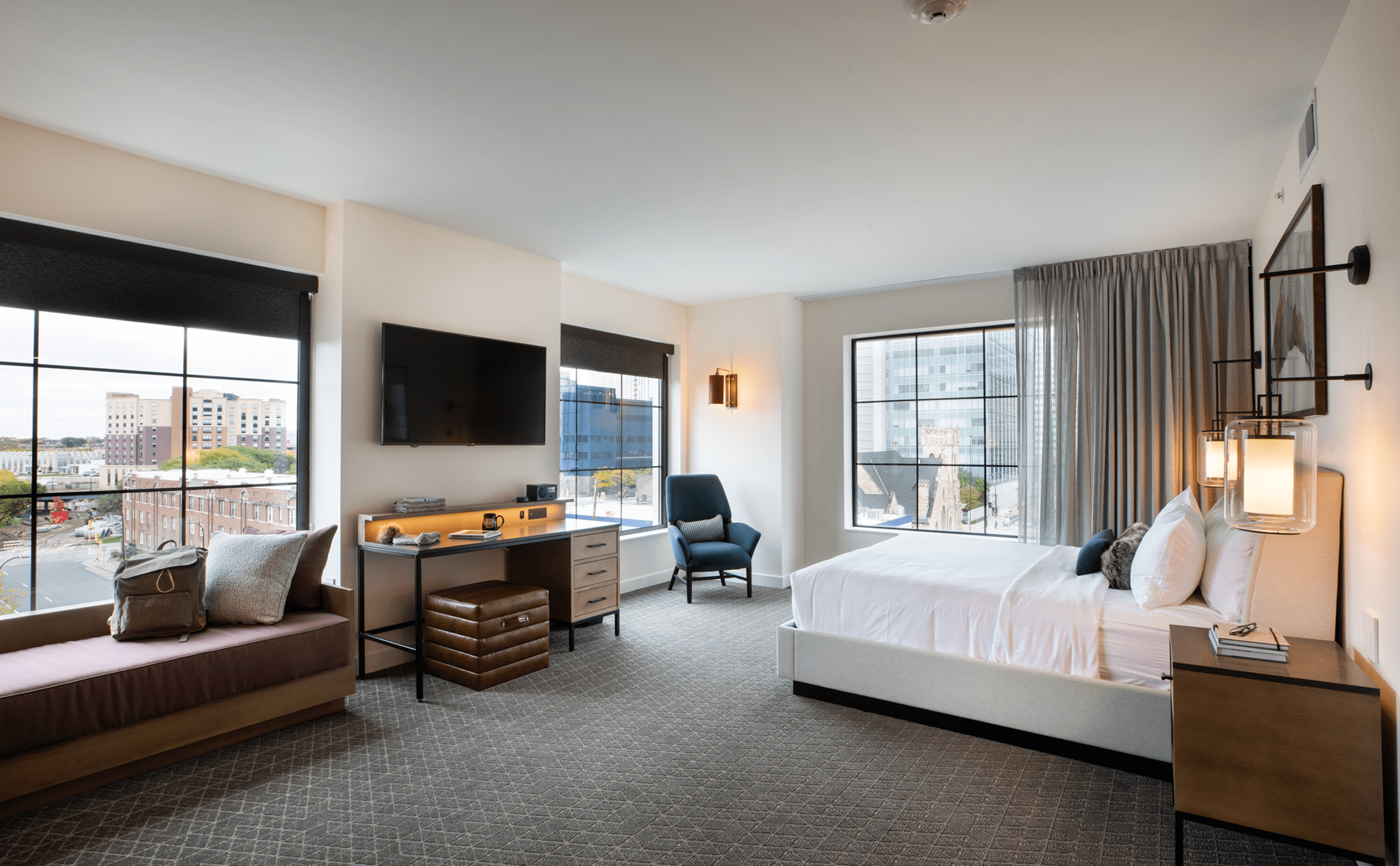 Modern hotel room with a large bed, desk, bench, armchair, wall-mounted TV, and city views through large windows.