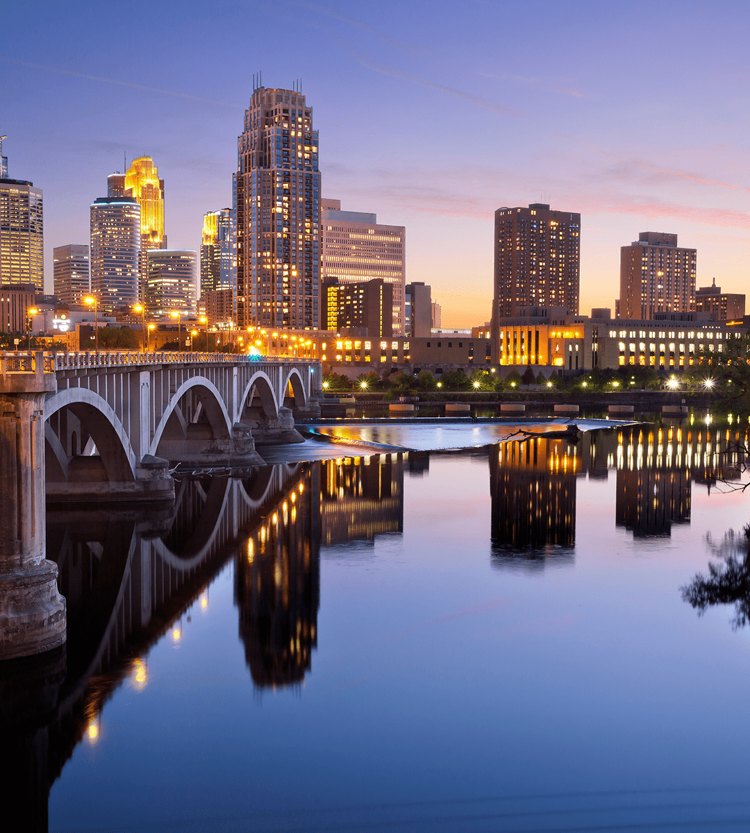 A city skyline at sunset with modern buildings, a bridge, and their reflections on a calm river—perfect for exploring new sights and discovering exciting things to do along the waterfront.
