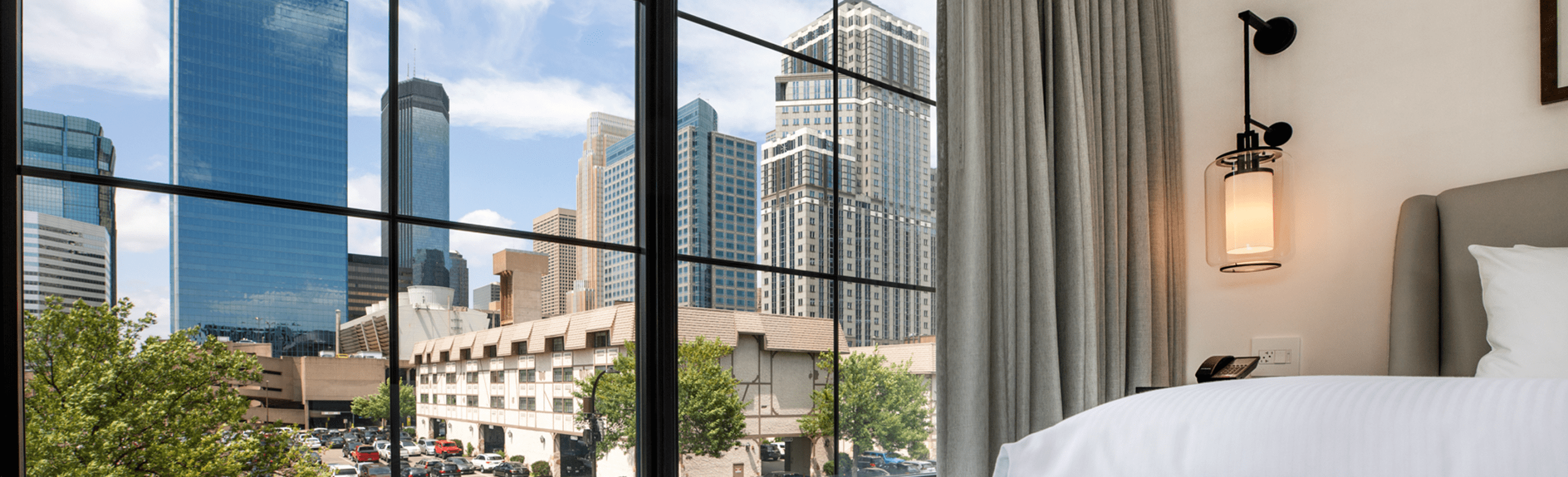 A hotel room with a bed and lamp, featuring a large window offering a clear view of downtown skyscrapers and a few trees outside.