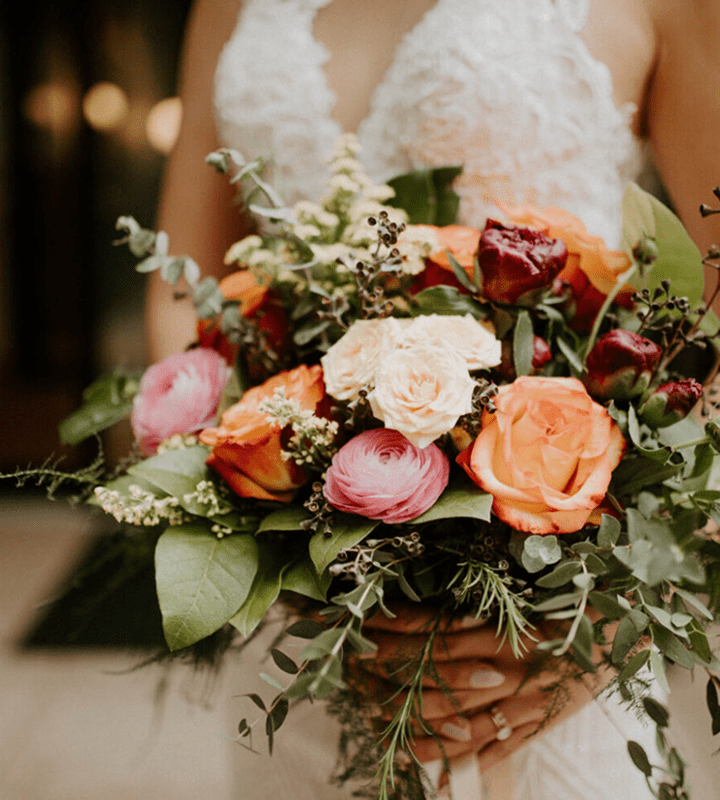 A person in a white lace dress holds a bouquet of roses and various flowers in shades of orange, pink, and cream, with green foliage.