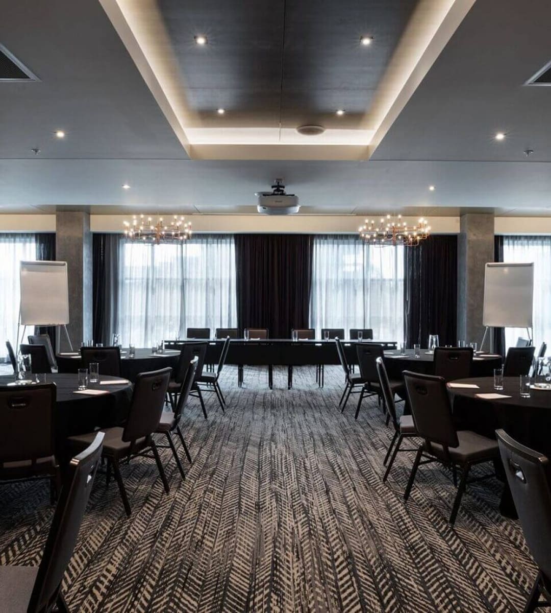 Conference room with round tables, chairs, flip charts, glasses, and a central rectangular table setup, under modern lighting and large windows with curtains.