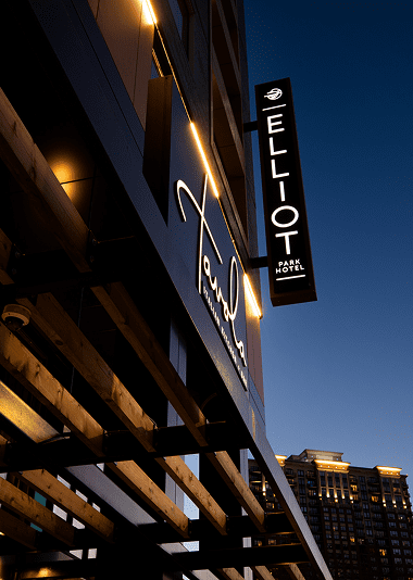 Exterior view of Elliot Park Hotel at dusk, featuring illuminated signage and wooden architectural elements with a city building in the background.