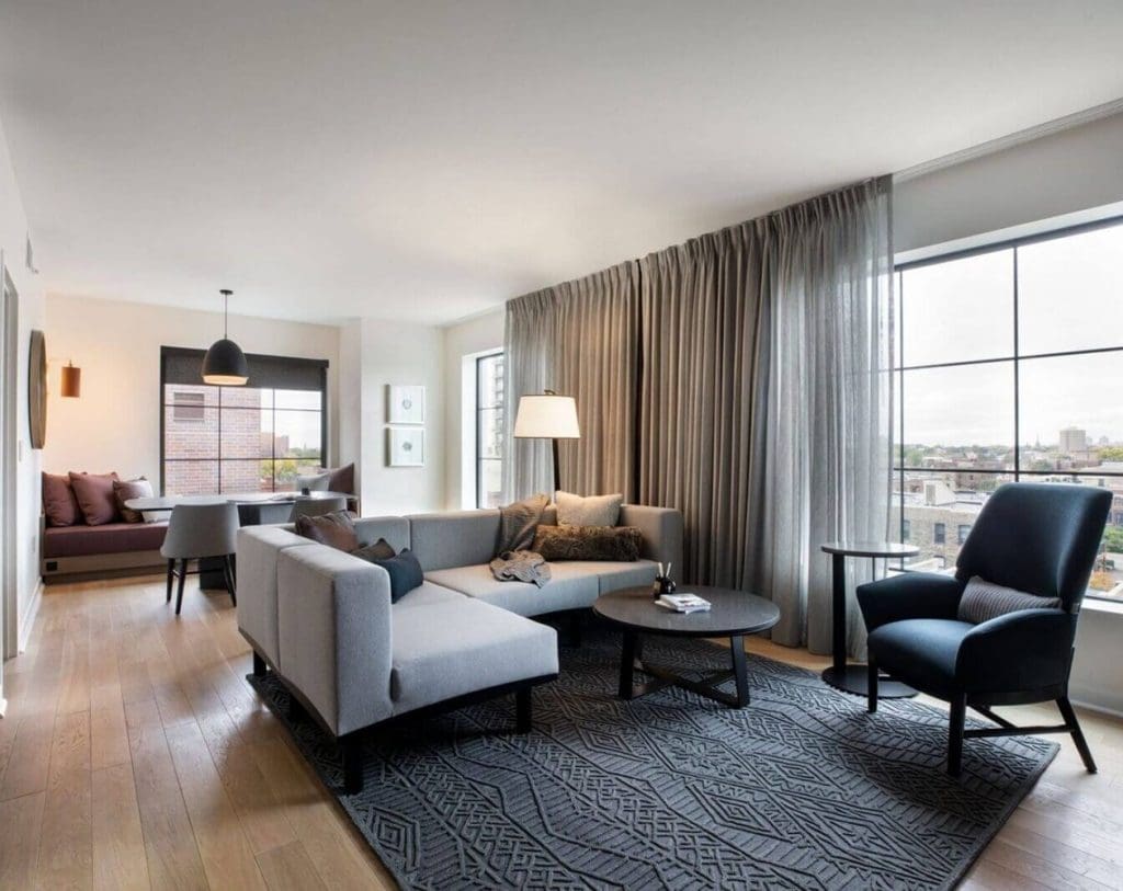 Modern living room with a gray sectional sofa, armchair, coffee tables, large windows with sheer curtains, and a window seat in the dining area.