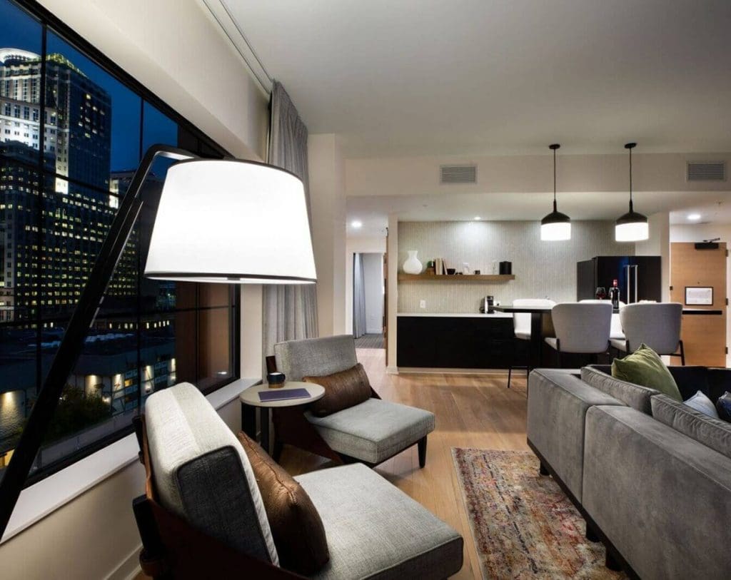 Modern apartment living room with large window overlooking city buildings, featuring gray chairs, sofa, floor lamp, and open kitchen with pendant lights in the background.