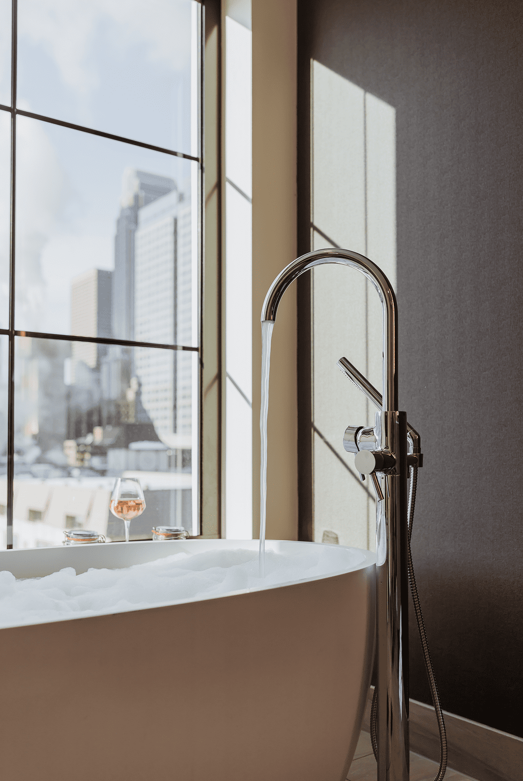 A modern bathtub with running water and bubbles sits by large windows overlooking a cityscape; a glass of wine and candles rest on the tubs edge.