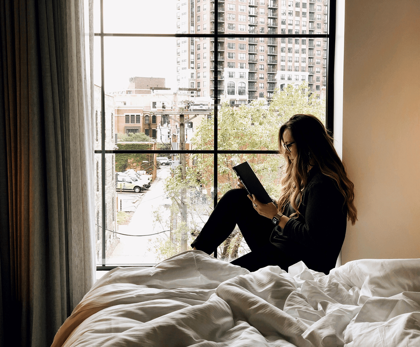 A person sits on a windowsill reading a book, with an urban cityscape visible outside. A bed with white blankets is in the foreground.