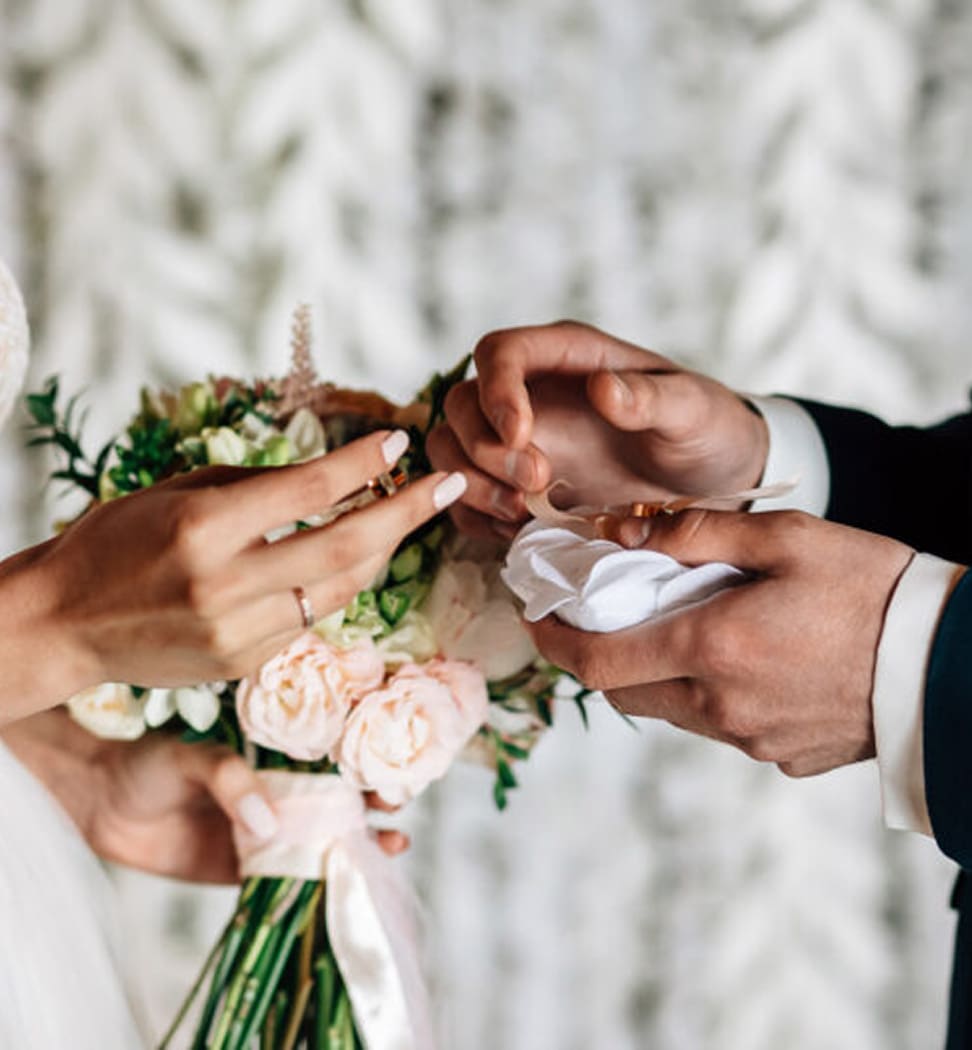 Close-up of a bride and groom exchanging wedding rings, with the bride holding a bouquet of white and pink flowers.