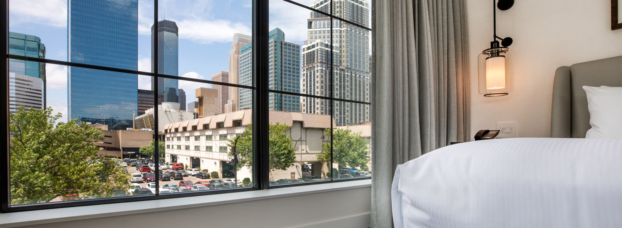 View of city skyscrapers through a large window from a hotel room with white bedding and a hanging wall lamp.