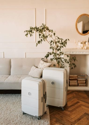 Two white suitcases on wheels are placed in a stylish living room with a beige sofa, a leafy plant, and a stack of books near a decorative mirror.