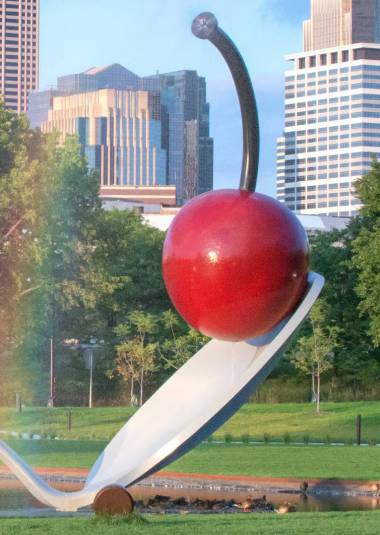 Large outdoor sculpture of a shiny red cherry balanced on a white spoon, set in a grassy park with city buildings in the background.