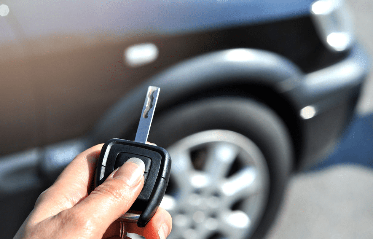 A hand holding a car key in front of a black car, with the car door and wheel visible in the background.