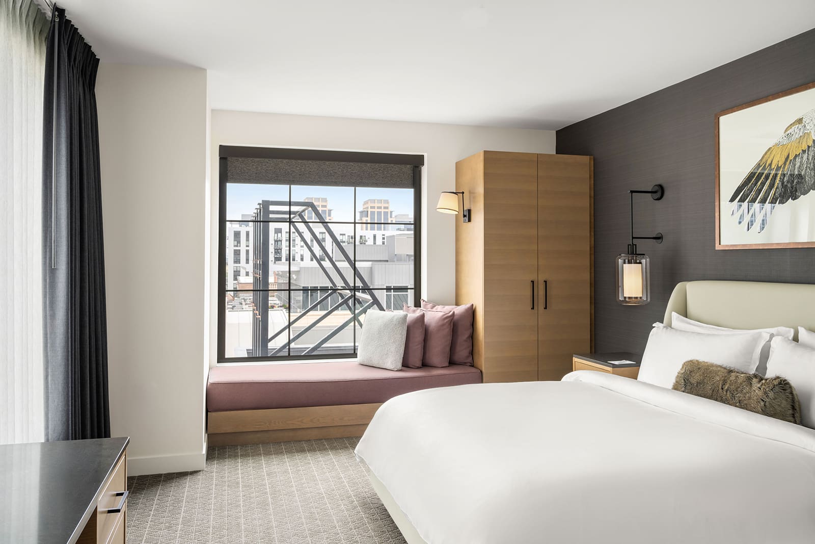 Modern hotel room with a large bed, wooden wardrobe, wall art, cushioned window seat, and city buildings visible through the window.