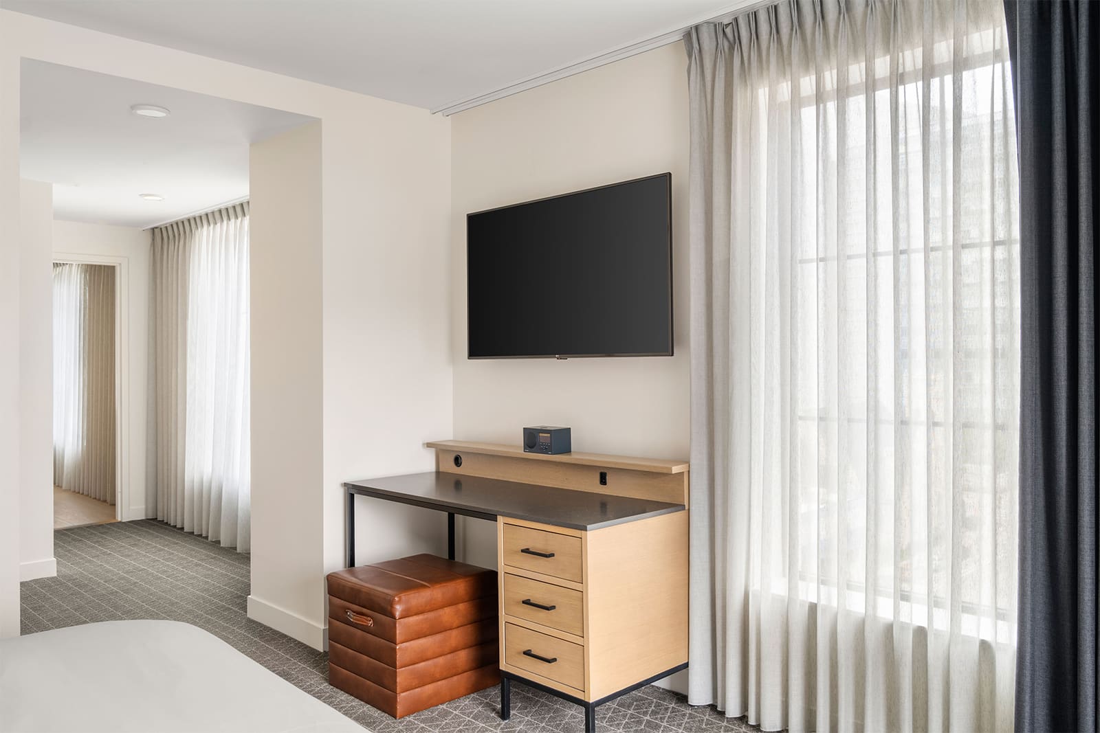 Modern hotel room with a wall-mounted TV, wooden desk, leather ottoman, and large window with sheer curtains letting in natural light.