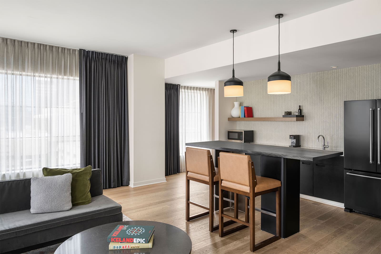 Modern kitchen and living area with black cabinets, island seating, gray sofa, wood floors, and large windows with sheer and dark curtains. Two pendant lights hang above the island.
