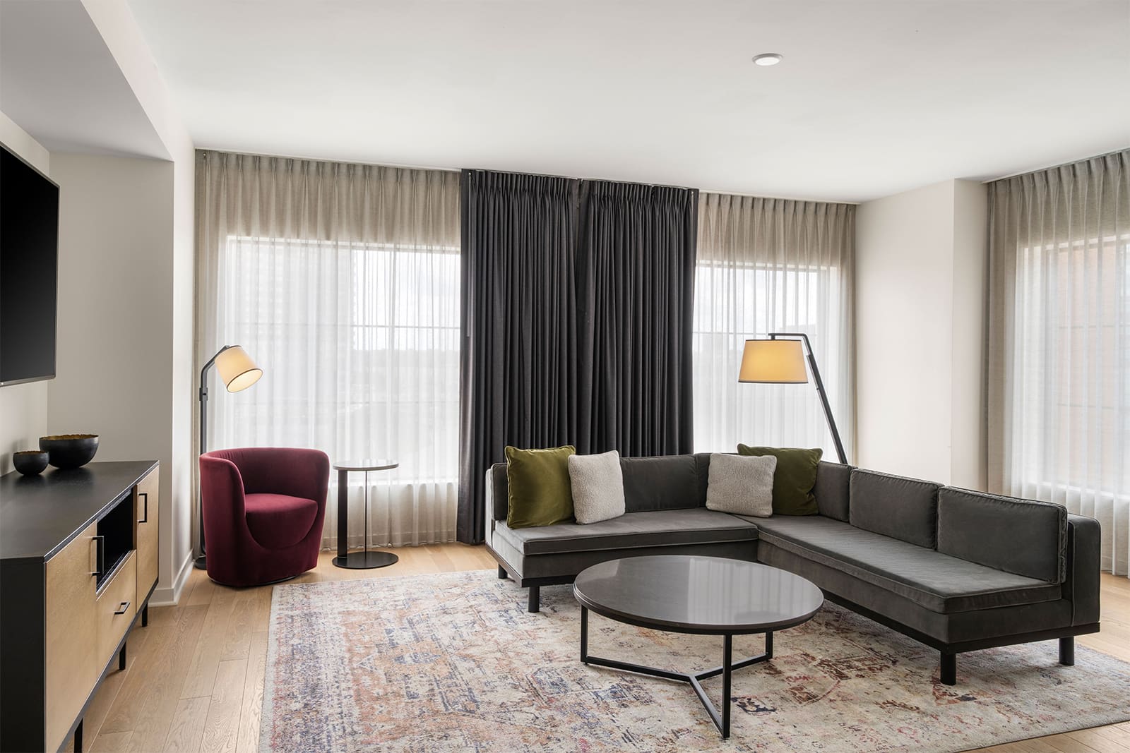 Modern living room with a gray sectional sofa, round coffee table, floor lamps, red chair, patterned rug, and large windows with sheer and blackout curtains.