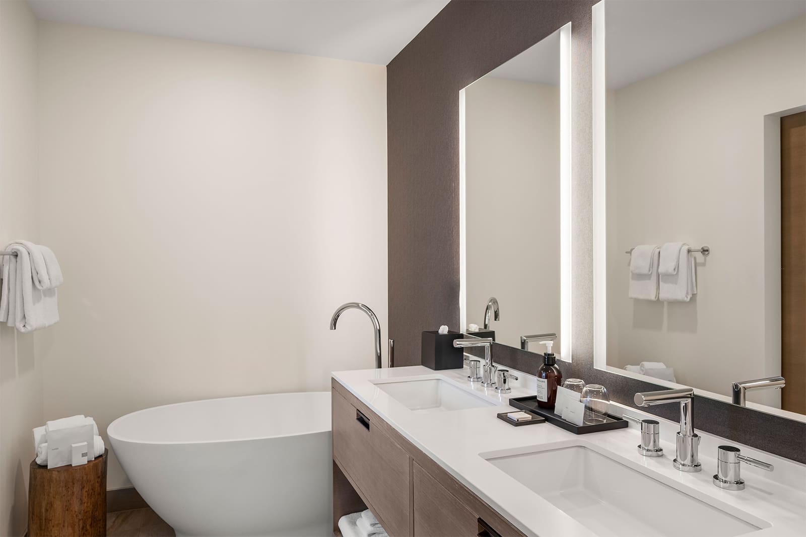 Modern bathroom with a freestanding bathtub, double sink vanity, wall-mounted mirror with lights, and towels neatly arranged on racks and a wooden stool.
