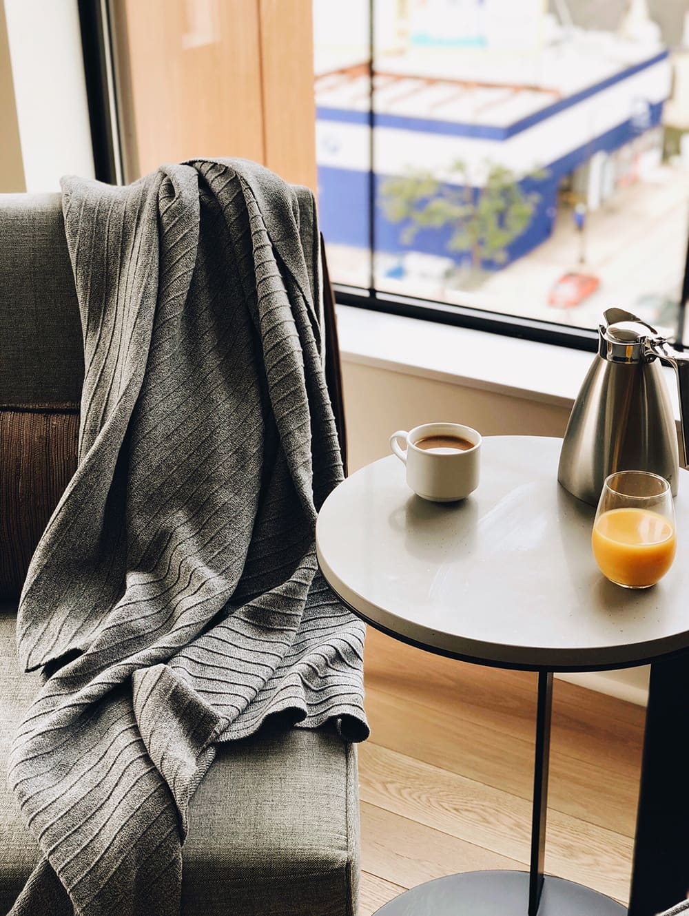 A gray blanket draped over a chair next to a round table with a cup of coffee, a glass of orange juice, and a metal carafe by a window.