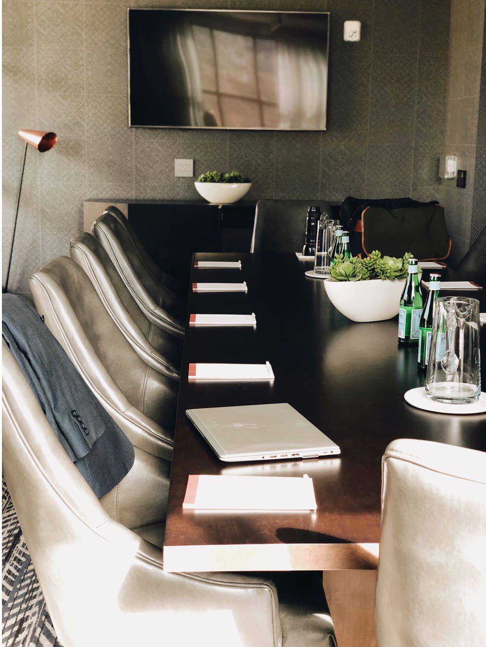 Conference room with a long table, leather chairs, notebooks, water bottles, a laptop, potted plants, and a wall-mounted TV. Sunlight streams through an unseen window.