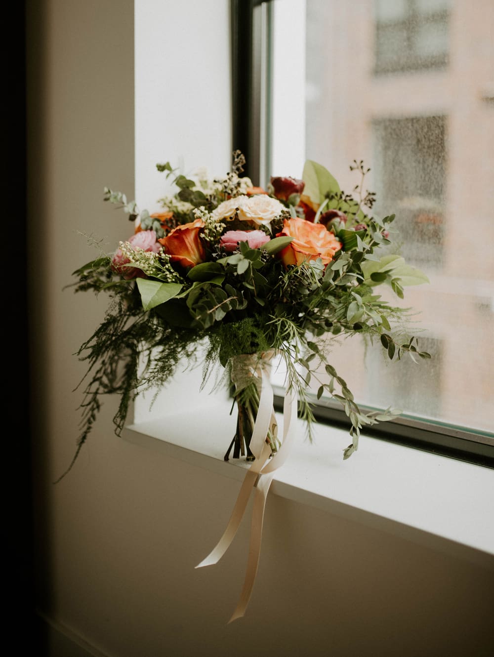 A bouquet of orange, pink, and white flowers with greenery and ribbons rests on a windowsill beside a rain-speckled window.