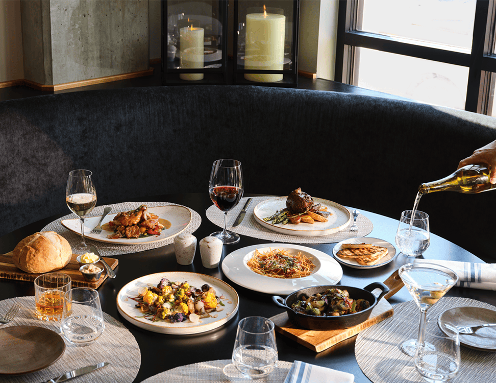 A round table set with assorted plated dishes, bread, wine glasses, and candles; someone pours white wine into a glass.